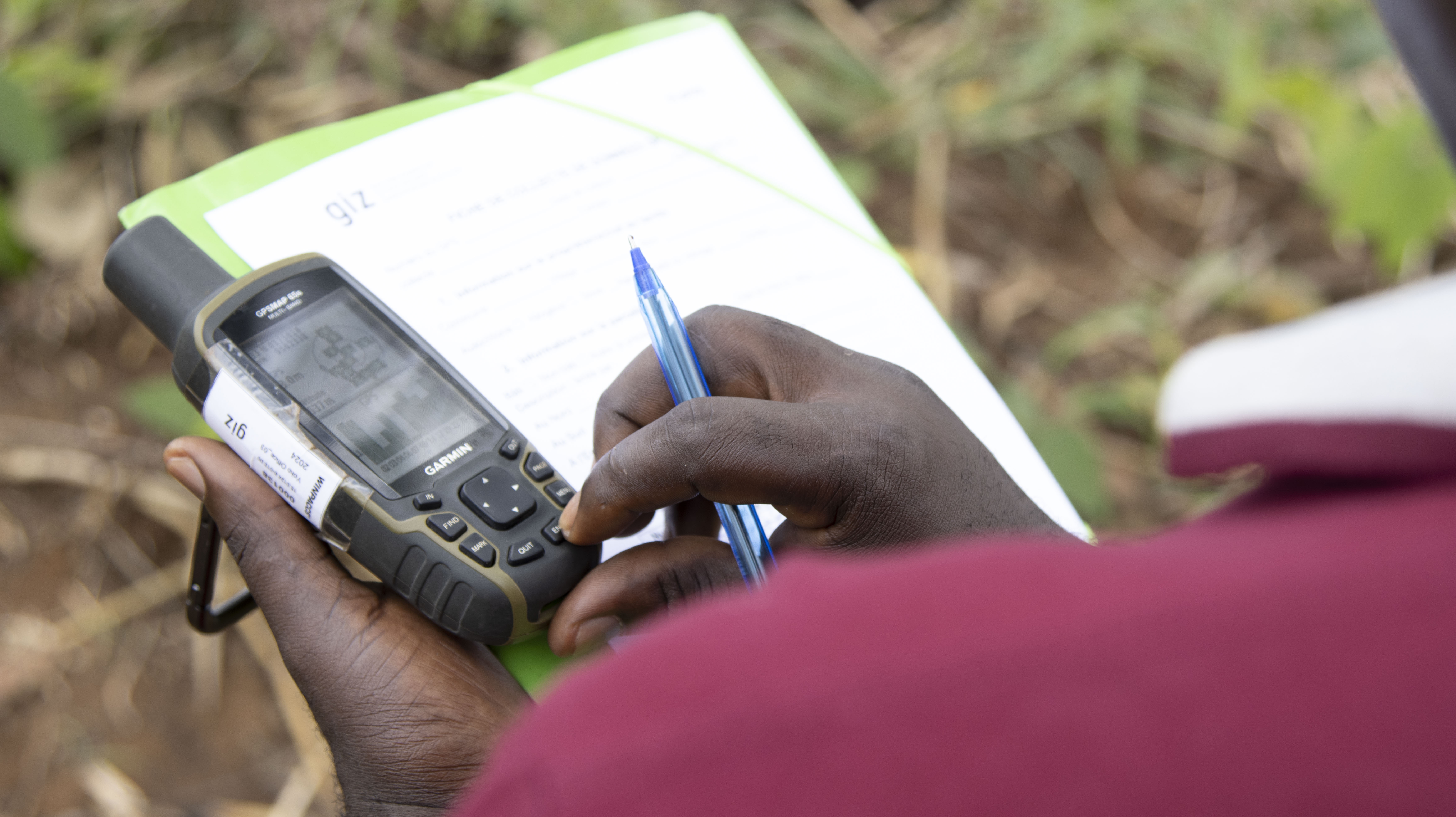 View over the shoulder, right hand holds a pen over a clipboard, left had holds a GPS tracking device.