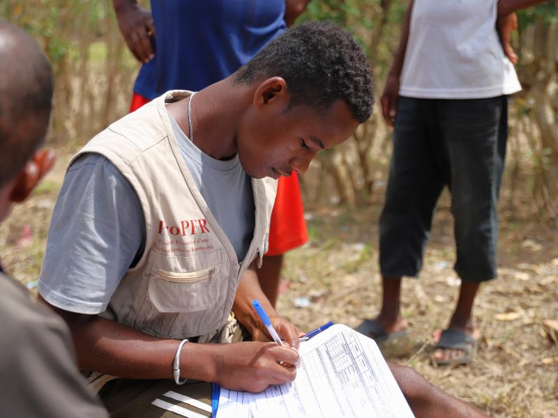 A man write on a sheet of paper, other people stand around him.